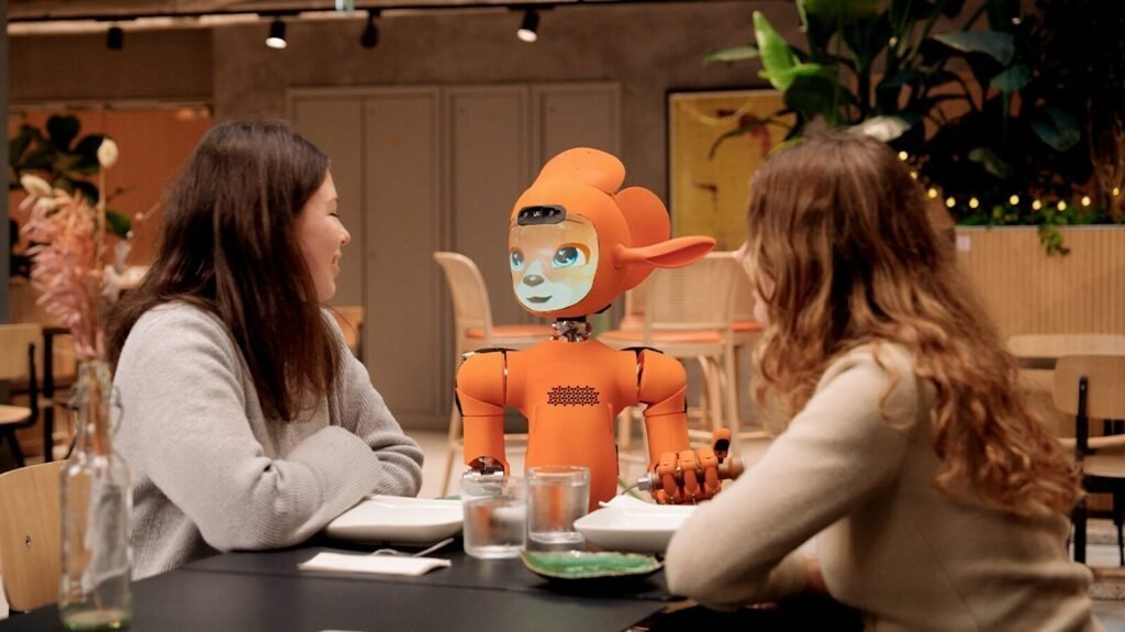 Two Women Talk To An Orange Robot At A Table.